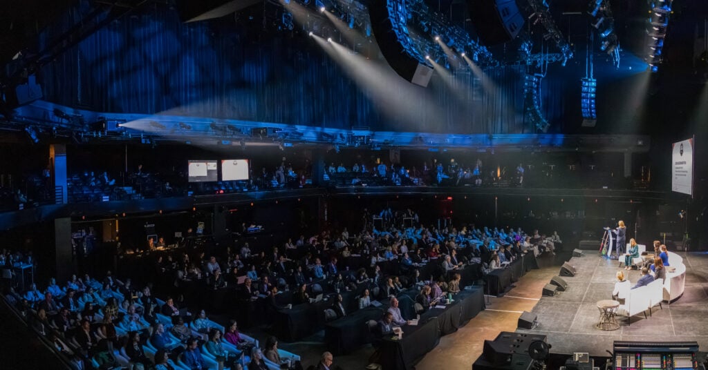 Wide view of a packed theater audience under blue lighting, facing a bright stage with seated speakers, dramatic spotlights, and event screens.