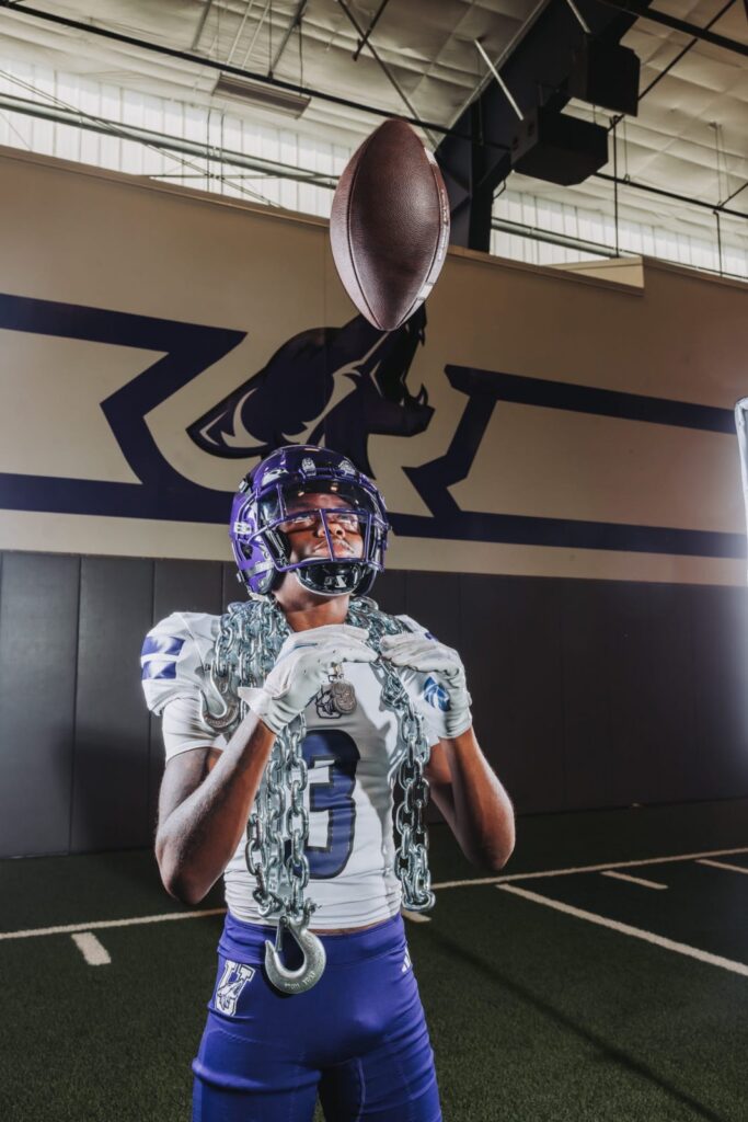 Football player in a white jersey and purple helmet catching a ball overhead indoors