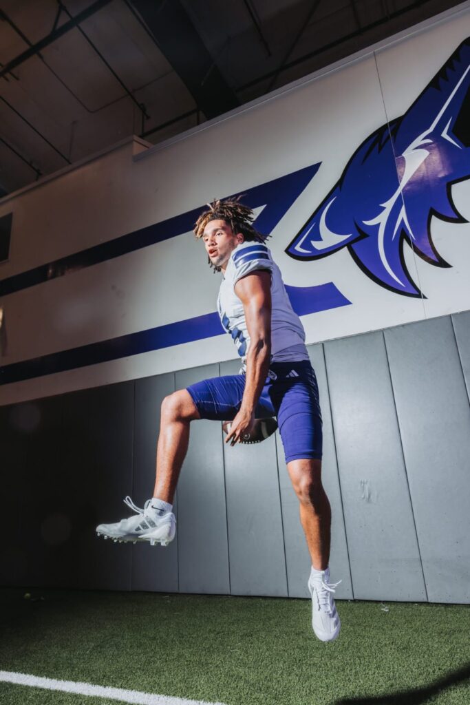 Football player in a white jersey jumping off a padded wall in an indoor facility