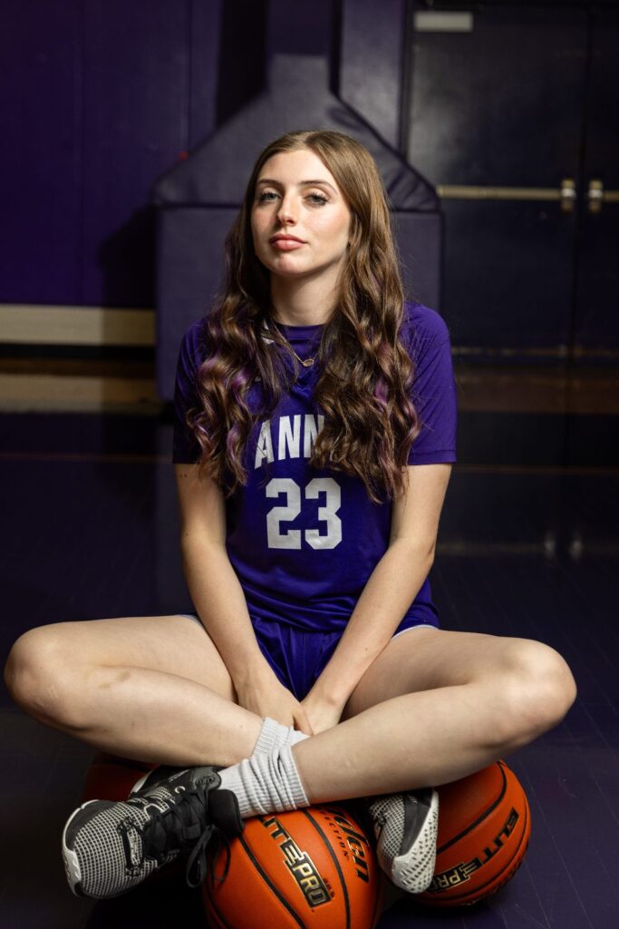 Girls basketball player in a purple number 23 jersey sitting cross-legged with basketballs