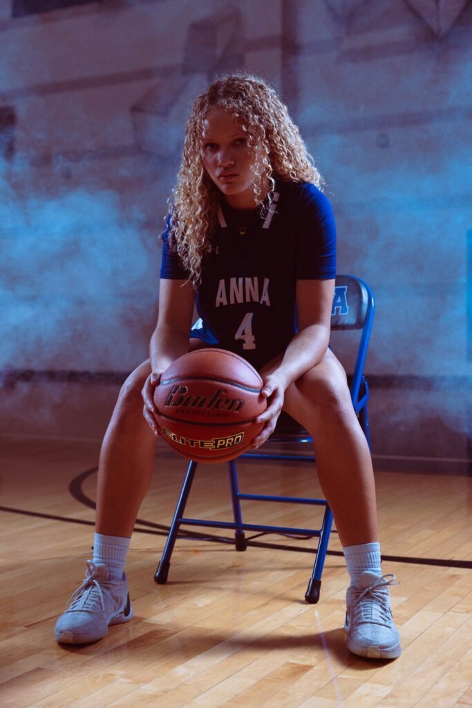 Girls basketball player in a purple jersey seated on a chair holding a basketball in a smoky studio