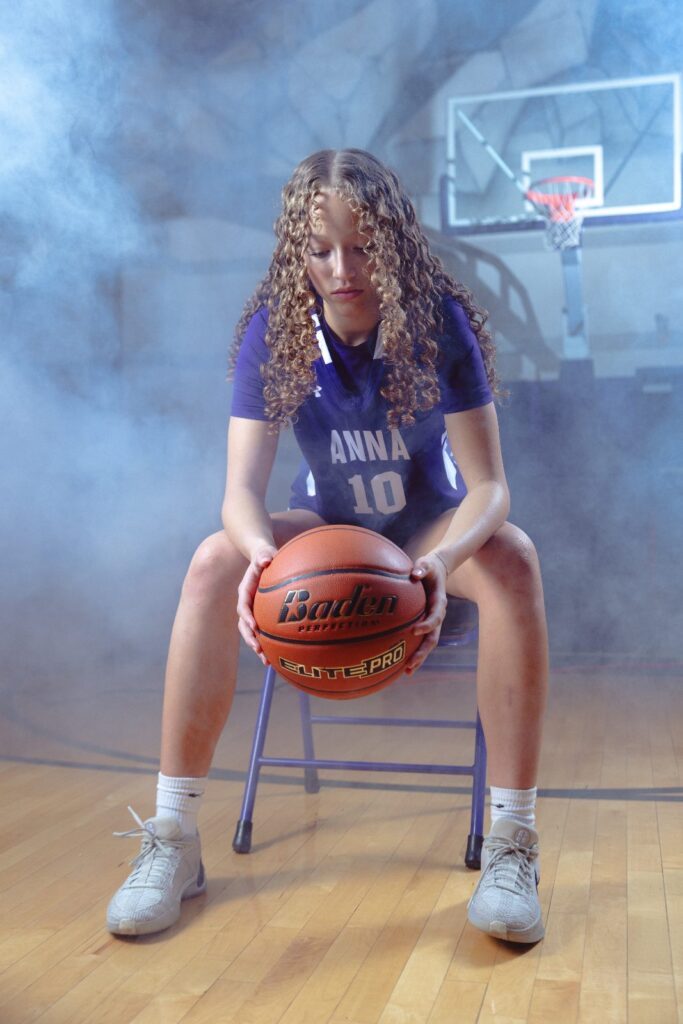 Girls basketball player seated with a basketball under a hoop in a hazy studio